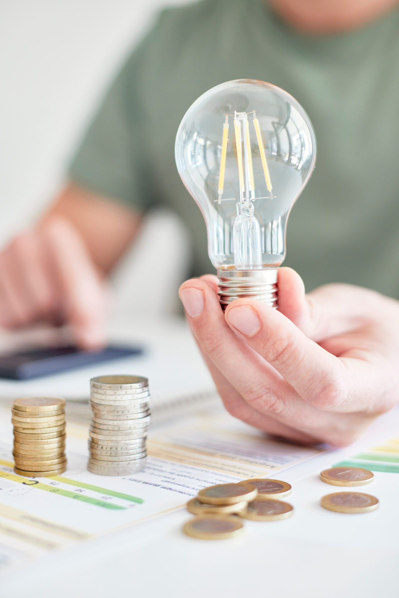 Jetzt Energieausweis Bielefeld sichern – Kosten sparen, Energieeffizienz prüfen und Umwelt schützen! Hand hält eine Glühbirne, daneben gestapelte Münzen und ein Energieausweis – Symbol für Energieberatung Bielefeld und Energieeffizienz in Bielefeld.