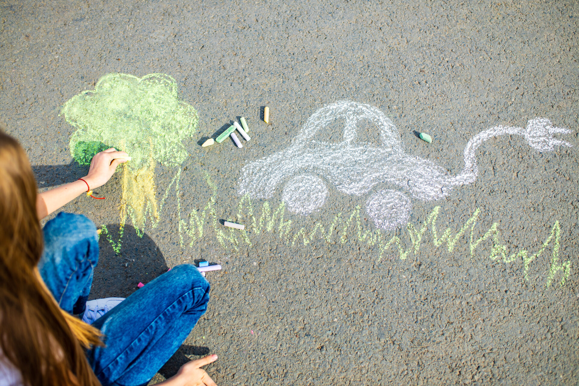 Bunte Träume von einer grünen Welt – Kinderzeichnung mit E-Auto und Baum Ein Kind sitzt auf dem Boden und malt mit bunter Kreide auf den Asphalt. Zu sehen sind eine grüne Baumkrone mit braunem Stamm, grasartige grüne Linien und ein weißes Auto mit einem Kabel und Stecker, das ein Elektroauto symbolisiert. Die Szene vermittelt den Zusammenhang zwischen Elektromobilität und Umweltschutz.
